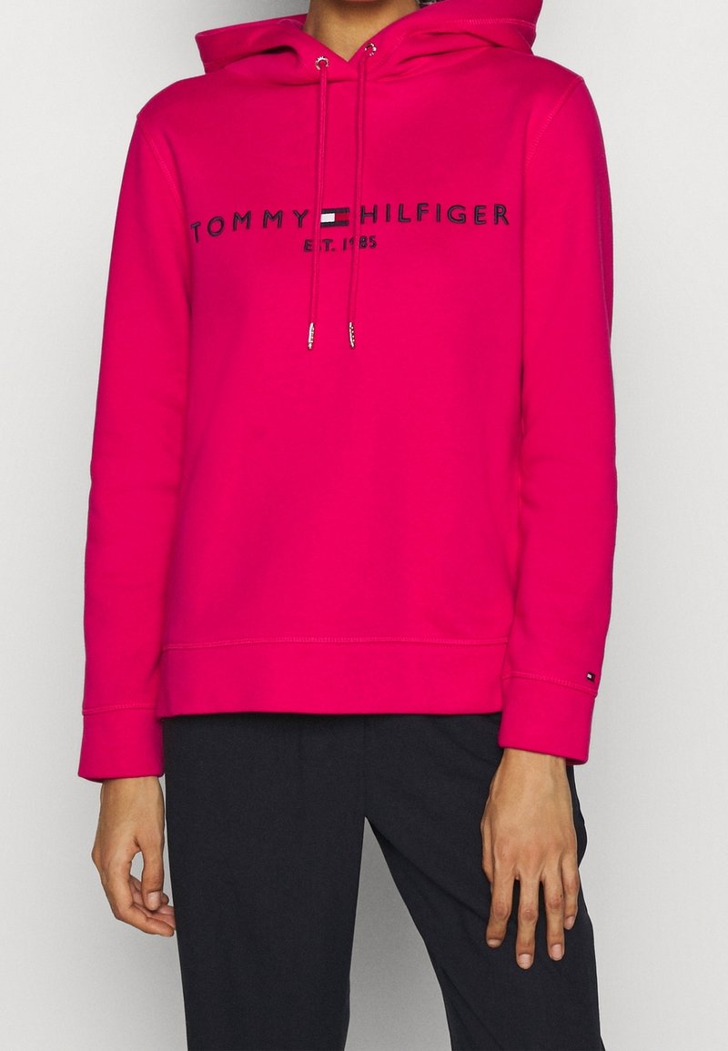 Personne portant un sweat à capuche Tommy Hilfiger rose vif avec logo et un pantalon noir, debout devant un arrière-plan clair uni.