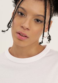 Ung person med fletninger iført gyldne hoops øreringe og hvid t-shirt med rund hals, der kigger let mod kameraet.