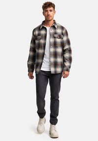 Camicia di flanella a quadri marrone, blu e crema; due tasche sul petto; abbinata a una t-shirt bianca e jeans scuri; stivali beige.