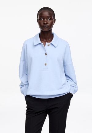 Kobieta ubrana w jasnoniebieską koszulę z długim rękawem, kołnierzykiem i trzema guzikami oraz czarne spodnie, stoi z rękami w kieszeniach na białym tle.