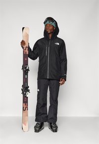 Schwarze wasserdichte Skijacke und Hose. Skischuhe mit schwarzen Akzenten. Hält Skier mit einer holzähnlichen Oberfläche und schwarzen Bindungen. Schwarze Goggles.