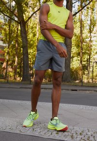 Helles gelbes ärmelloses Sportshirt, graue Shorts und neongrüne Laufschuhe mit roten Akzenten; im Freien zwischen Bäumen.