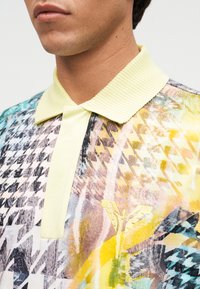 Άνδρας που φοράει πολύχρωμο, εμπριμέ πόλο πουκάμισο με ανοιχτόκίτρινο ριμπ λαιμό και κίτρινο κεντητό φτερωτό σχήμα στο στήθος.