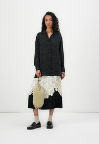 Schwarze gemusterte Hemdbluse mit langen Ärmeln, fließender cremefarbener, blumenbedruckter, plissierter Rock, beiger Tasche und schwarzen Schuhen. Strukturierter und geschichteter Look.