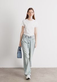 Camiseta blanca de manga corta, jeans azul claro y zapatillas blancas. Llevando un bolso azul, de pie contra un fondo simple.