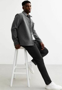Chaqueta de mezcla de lana gris con cuello y cierre de botones, combinada con pantalones negros y zapatillas blancas, sentado en un taburete blanco.