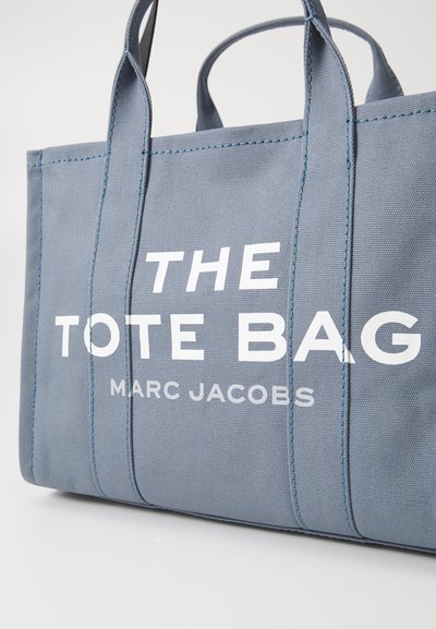 Pelēka kanvas iepirkumu soma ar divām rokām, uz kuras priekšpuses redzams drosmīgs balts teksts "IEPIRKUMU SOMA" un "MARC JACOBS". Redzami izšūti akcenti.