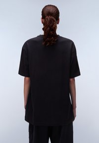 Schwarzes, oversized T-Shirt mit kurzen Ärmeln, aus Baumwolle. Besitzt einen runden Ausschnitt und einen geraden Saum, mit einem dezenten Markenlabel an der Seite.