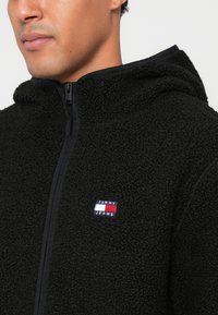 Mustavillainen fleece-huppari vetoketjulla, jossa on pieni brodeerattu "TOMMY JEANS" -logo. Pehmeä pinta, kontrastiväriset yksityiskohdat reunoilla.