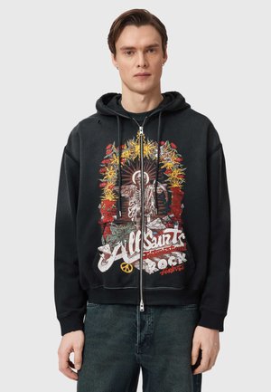 Junger Mann trägt schwarzen Kapuzenpullover mit Reißverschluss und buntem All Saints Rock Festival-Motiv sowie dunkle Jeans, steht vor grauem Hintergrund.