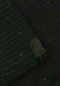 Donkergroen gebreid stof met gele spikkels, met een zwart label met gouden tekst "JACK & JONES." Geribbeld textuurontwerp.