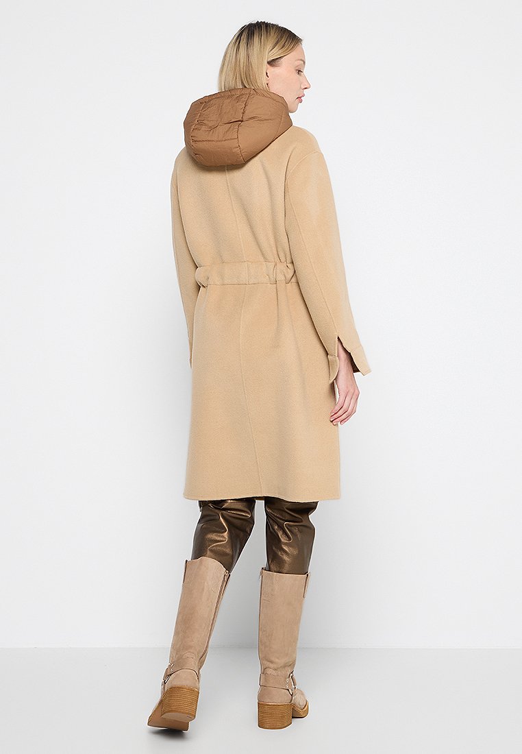 Abrigo de mezcla de lana beige con capucha marrón acolchada, cintura ceñida por un cinturón y mangas anchas, combinado con pantalones marrones brillantes y botas beige de caña alta.