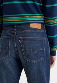 Personne portant un jean en denim bleu avec des coutures orange et un pull rayé bleu marine vue de dos au niveau de la taille.