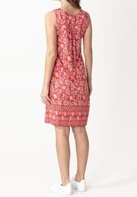 Femme portant une robe rouge sans manches avec un motif floral blanc et une bordure, associée à des baskets blanches, debout de dos.