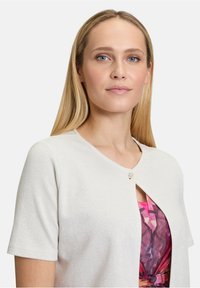 Lichtgrijze cardigan met korte mouwen, subtiele glans en ronde hals. Bevat een decoratieve knoopsluiting aan de voorkant.