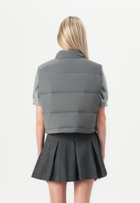 Donna bionda di spalle che indossa un gilet imbottito grigio, una maglietta a maniche corte e una gonna pieghettata grigio scuro su uno sfondo bianco.