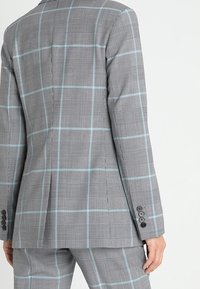 Grå rutiga kavaj med blå linjer, strukturerad design och texturerad tyg. Fem-knappliknande ärmslut och riven ryggdetalj.
