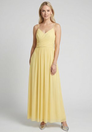 Blonde vrouw in een lange, vloeiende gele mouwloze jurk met spaghettibandjes, staand tegen een effen lichtgrijze achtergrond.