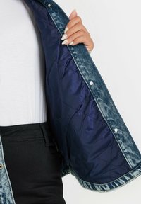 Jeansjacka med en ljusblå ytterdel och en mörkblå kviltad foder. Har tryckknappar och en något fransad fåll.