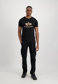 Schwarzes Baumwoll-T-Shirt mit goldfarbigem Grafiklogo. Getragen mit schwarzen Cargo-Hosen und schwarzen Schuhen. Einfaches Rundhalsausschnitt-Design.