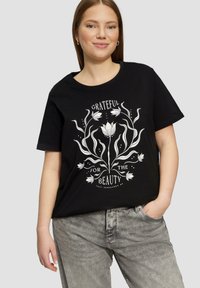 T-shirt en coton noir arborant un design graphique textuel floral blanc portant l'inscription « RECONNAISSANT POUR LA BEAUTÉ » entouré de lignes et de points.