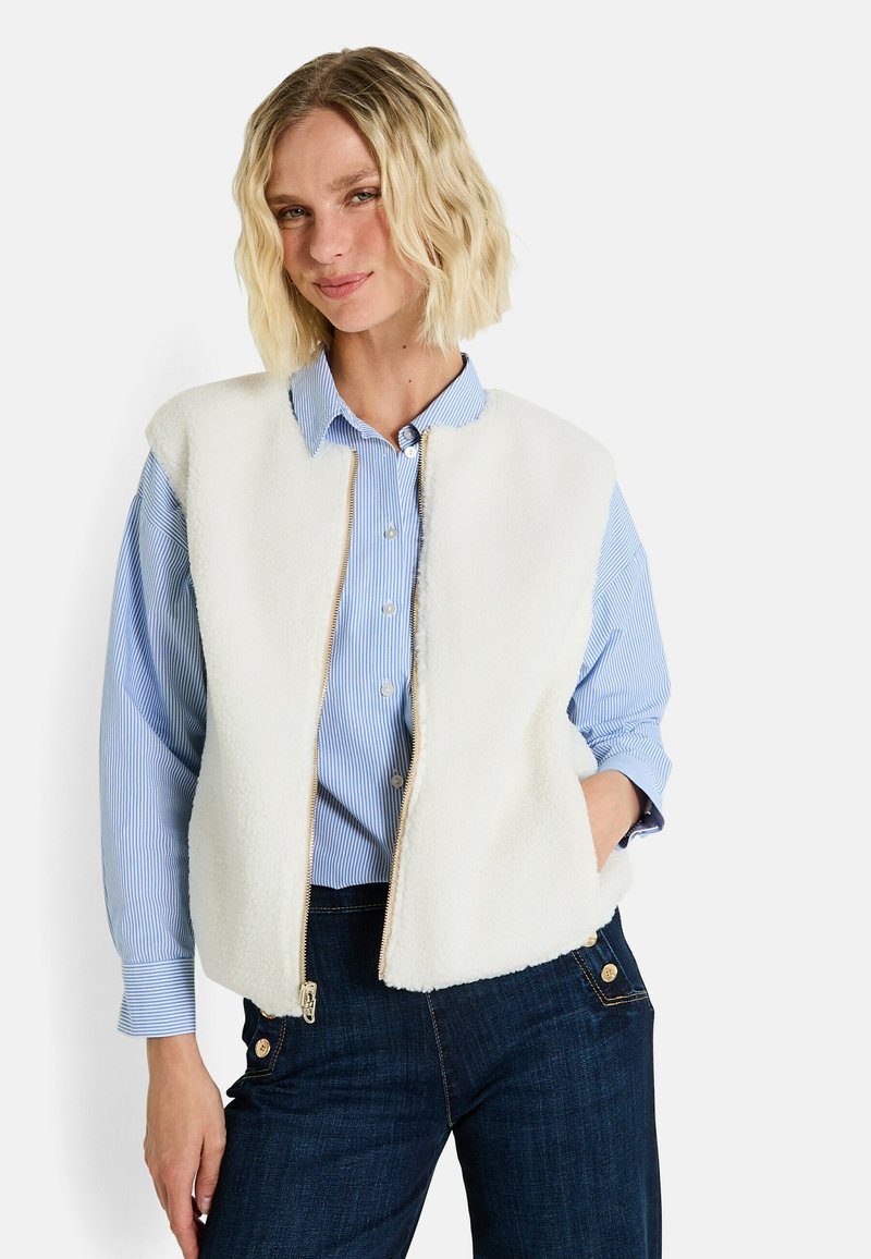 Gilet en polaire blanche avec fermeture éclair, porté sur une chemise à boutons à rayures bleu clair. Assorti avec un pantalon en denim foncé.