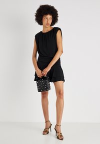 Vestido preto com mangas curtas bufantes e cintura franzida, com barra em camadas de babados. Modelo segurando uma pequena bolsa preta com tachas prateadas e usando saltos com tiras.