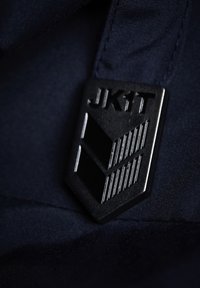 Svart metallbadge med texten "JK1T", som har chevronmönster och parallella linjer, fäst vid en mörk marinblå tygbakgrund.