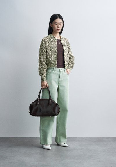 Femme portant un cardigan beige texturé, un top marron, un pantalon ample vert clair, des talons blancs, tenant un grand sac à main marron foncé, debout sur un sol gris.