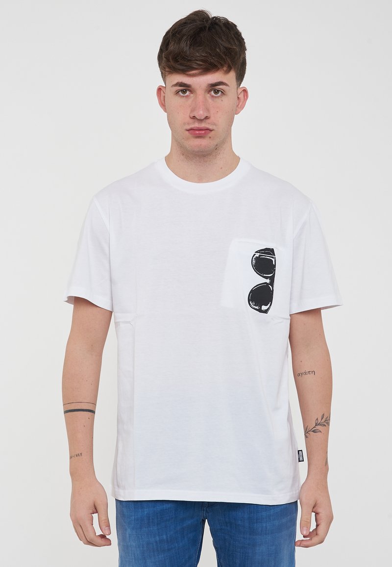 T-shirt bianca in cotone con maniche corte, dotata di una tasca sul petto e una grafica di occhiali da sole neri. La vestibilità è rilassata. Visibili jeans in denim.