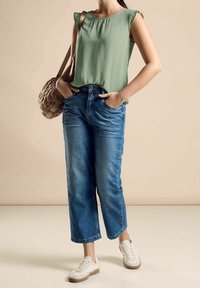 Haut vert sans manches avec un col froncé, associé à un jean bleu à jambes larges et des baskets blanches. Sac à main tressé marron en accessoire.