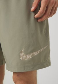 Pantaloni a bermuda Nike color khaki con un logo testurizzato e a motivi in beige chiaro. Il tessuto è liscio con un taglio sartoriale. Mano posizionata sul tessuto.