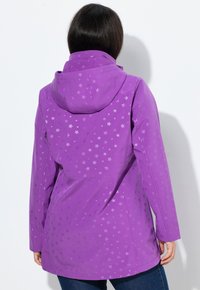 Nainen, jolla on päällä violetti huppari, jossa on hienovarainen tähkäkuvio, näkyy selältä yksinkertaista valkoista taustaa vasten.