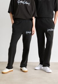 To personer iført svarte matchende joggebukser og t-skjorter med hvit logo-tekst, stående med hendene i lommene og hvite sneakers.
