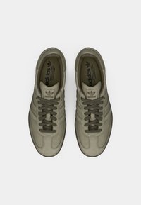 Šedé tenisky Adidas s hladkým koženým svrškem, kulatou špičkou, silnou gumovou podrážkou a designem se třemi pruhy, vybavené olivově zelenými tkaničkami a logem.