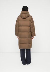 Hnědý péřový kabát s kapucí, s horizontálním prošíváním, střední délky po lýtka a hladkým povrchem. Pohled zezadu ukazuje detaily loga na límci.