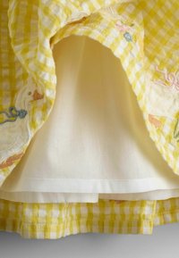 Dzeltens un balts ginghama audums ar izšūtiem baltiem pīļu un ziedu motīviem, rādot iekšpusi, kas oderēta ar vienkāršu baltu audumu.