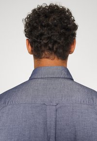 Camisa de mezclilla azul para hombre con cuello puntiagudo, detalle de canesú y costuras visibles. La tela tiene una textura suave y un color uniforme.
