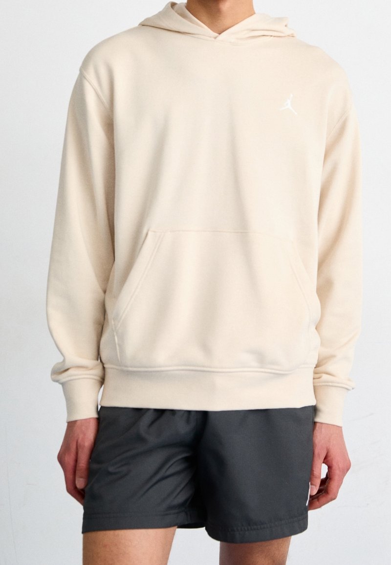 Cream hooded sweatshirt with a front pocket, ribbed cuffs, and a small white logo on the left chest, paired with dark shorts.