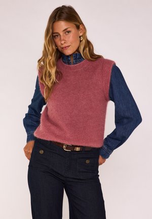 Frau trägt einen pinken, gestrickten ärmellosen Pullover über einem blauen Jeanshemd und dunklen Jeans, posiert mit den Händen in den Hüften vor einem einfarbigen Hintergrund.