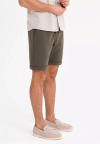 Mäns olivgröna shorts med en texturerad tyg, vikta fållar och sidofickor, parat med krämfärgade slip-on skor på barfota.