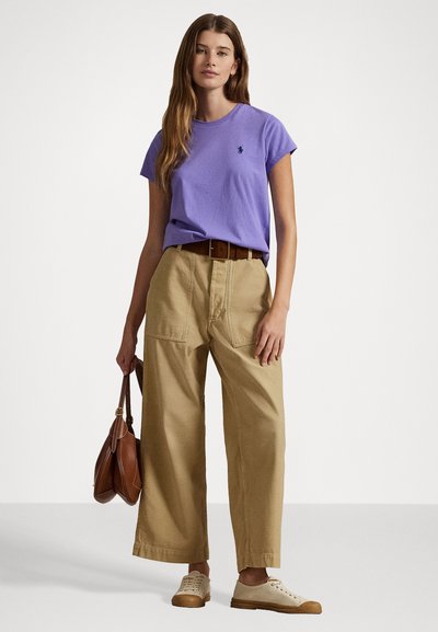 T-shirt di cotone viola con maniche corte, abbinato a pantaloni larghi kaki. Una cintura in pelle marrone e sneakers color crema completano il look.