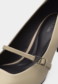 Μπεζ γυναικείο παπούτσι Mary Jane με λεπτό λουράκι και διακοσμητική γεωμετρική αγκράφα σε χρυσό τόνο, με μαύρο επενδυμένο εσωτερικό.