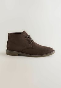 Botas de tobillo de gamuza marrón con cordones, con punta redondeada y costuras en contraste en la suela, exhibidas sobre un fondo claro.