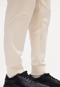 Cremfarbene Sweatpants mit strukturiertem Finish und Nahtdetails, kombiniert mit schwarzen Sneakers, die eine glatte Oberfläche und Schnürsenkel aufweisen.