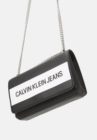 Černá texturovaná kabelka z umělé kůže se stříbrným řetízkem a bílou kapsou s nápisem "CALVIN KLEIN JEANS." Obdélníkový tvar.