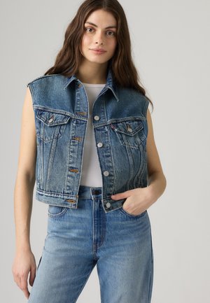 Junge Frau mit langen braunen Haaren trägt eine ärmellose blaue Denimjacke und hellblaue Jeans, sie steht vor einem einfarbigen hellen Hintergrund.