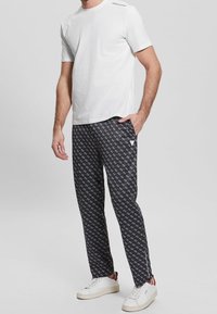 Muž v bílém krátkém rukávu, s vzorovanými černými kalhotami s malými bílými logy, v bílých teniskách, stojící s jednou rukou v kapse.