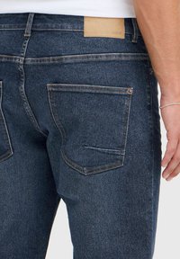 Jeans in denim blu scuro caratterizzati da una superficie liscia, tasche posteriori con dettagli di cucitura e un'etichetta in pelle marrone chiaro sulla vita.
