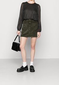 Blusa a righe nere con maniche a sbuffo, abbinata a una gonna in velluto a coste verde con bottoni frontali e tasche. Accessoriata con una borsa nera.